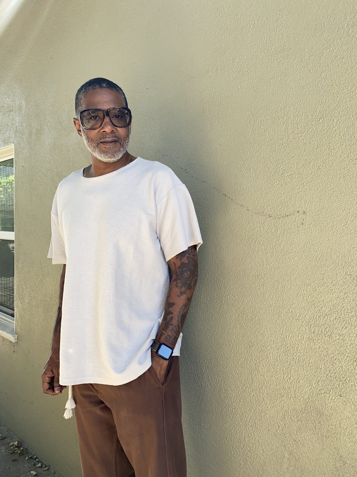 Miguel Collins standing against a warm stucco wall, showing a tattoo sleeve